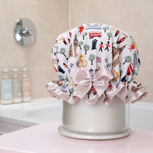 May include: A white shower cap adorned with a London-themed pattern, including red buses and iconic buildings. It features a pink satin bow and trim. The cap is displayed on a white container.