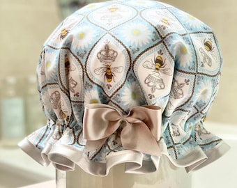 May include: A light blue shower cap with a repeating pattern of bees, crowns, and daisies. The cap has a ruffled edge and a large, light brown satin bow. The design is a mix of floral and insect motifs.