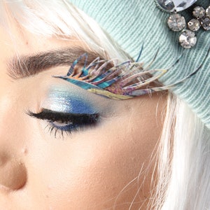 Può includere: Primo piano di un occhio di donna con ombretto blu e argento e un design a forma di piuma colorato sopra l'occhio. La donna indossa un berretto blu chiaro con un abbellimento di strass.