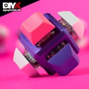 May include: A purple, white and pink mechanical keyboard keycap set. The keycaps are arranged in a cube shape, with each side showing a different colour. The keycaps are made of plastic and have a smooth, matt finish.