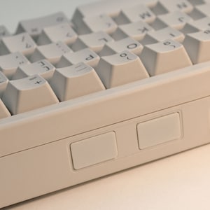 May include: A white keyboard with a beige case. The keyboard has a standard layout with keys for letters, numbers, and symbols. There are two additional buttons on the right side of the keyboard.