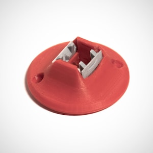 May include: A red 3D printed base with a gray plastic clip. The base has a circular shape and a small hole on the side. The clip is designed to hold something in place.