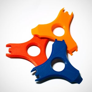 May include: Three 3D-printed tools in orange, red, and blue. Each tool has a unique shape with a central hole and a forked end. The tools are arranged on a white surface, showcasing their design and color variations.