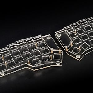Könnte beinhalten: Eine geteilte Tastatur aus transparentem Acryl mit Aussparungen für die Tastenkappen. Die Tastatur besteht aus zwei separaten Teilen, die durch kleine Metallabstandshalter verbunden sind. Das Design ist minimalistisch und modern, mit Fokus auf Transparenz und Funktionalität. Die Tastatur liegt auf einer schwarzen Oberfläche.