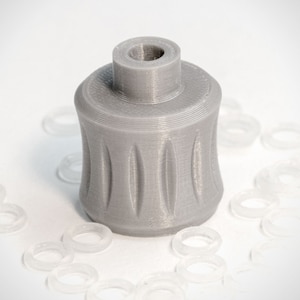 Puede incluir: Un tapón de botella gris impreso en 3D con un diseño texturizado. El tapón está hecho de plástico y tiene una parte superior redonda.