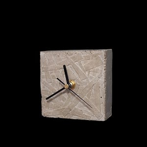 May include: A square concrete clock with black hands and a gold-colored center. The clock's face has a textured, mosaic-like pattern. The clock is a modern, minimalist design.
