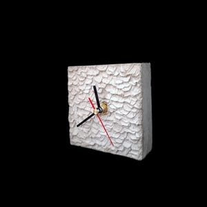 May include: A square, textured white clock with black and red hands. The clock has a gold-colored center and a unique, layered surface design. The clock is a decorative item.