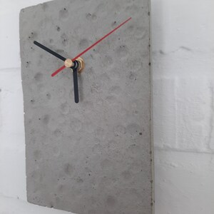 May include: A grey concrete clock with black hands and a red second hand. The clock is mounted on a white wall.