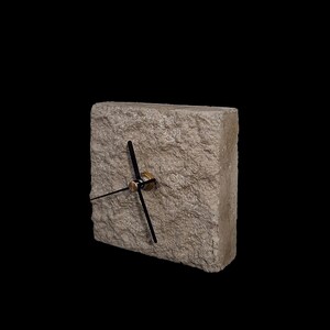 May include: A square clock made of textured, light-colored stone. The clock has black hands and a gold-colored center. The clock is set against a black background.