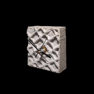 May include: A square, textured concrete clock with a waffle-like pattern. The clock has black hands and a gold-colored center. The clock is a light gray color and sits against a black background.