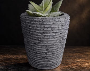 May include: A gray, tapered cylindrical planter with a textured, horizontally ribbed surface. The planter contains a small, light green succulent plant with pointed, fleshy leaves that extend slightly over the top rim.