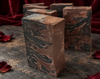 May include: Several rectangular bars of soap featuring a marbled pattern of dark charcoal gray and muted brown tones. The soap bars have a smooth, solid texture with visible swirls throughout the surface, arranged in a cluster on a dark surface.