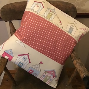 May include: A cream and red patterned throw pillow with a design of colorful beach houses. The pillow has a red background with white polka dots and a cream border with a row of beach houses.