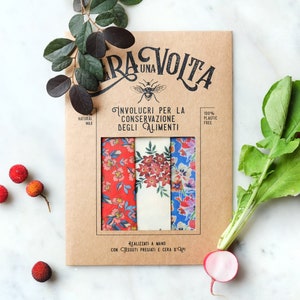 Può includere: Un set di tre involucri per alimenti riutilizzabili con motivi floreali. Gli involucri sono realizzati con cera d'api, cotone biologico e resina d'albero. La confezione è in cartone marrone con il testo "Era Una Volta" e "Involcri per la conservazione degli alimenti" in nero.