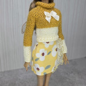 May include: A fashion doll with long brown hair wearing a mustard yellow and cream knit sweater with a white satin bow at the neck. The doll wears a yellow skirt featuring a white floral pattern with dark red centers and green leaves, accented by a ruffled white trim along the side.