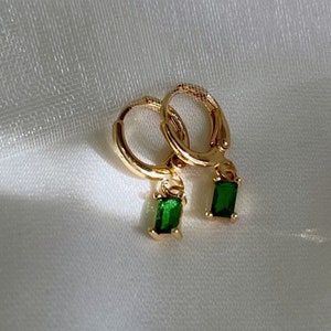 May include: A pair of gold-colored hoop earrings with small, rectangular emerald green gemstones dangling from the bottom. The earrings are displayed on a white, textured fabric.