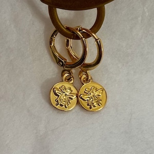 May include: A pair of gold-colored hoop earrings with a small, round charm. Each charm features a detailed embossed bee design. The earrings are suspended from a gold-colored hook.