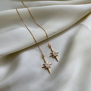 May include: A pair of gold-tone threader earrings featuring a starburst design with a central oval-shaped clear stone and small accent stones at the points of the star, suspended from delicate gold-tone box chains.