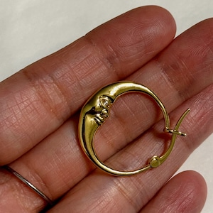 以下が含まれることがあります： 顔のデザインが施された金の三日月形のフープイヤリング。イヤリングは手で持たれています。