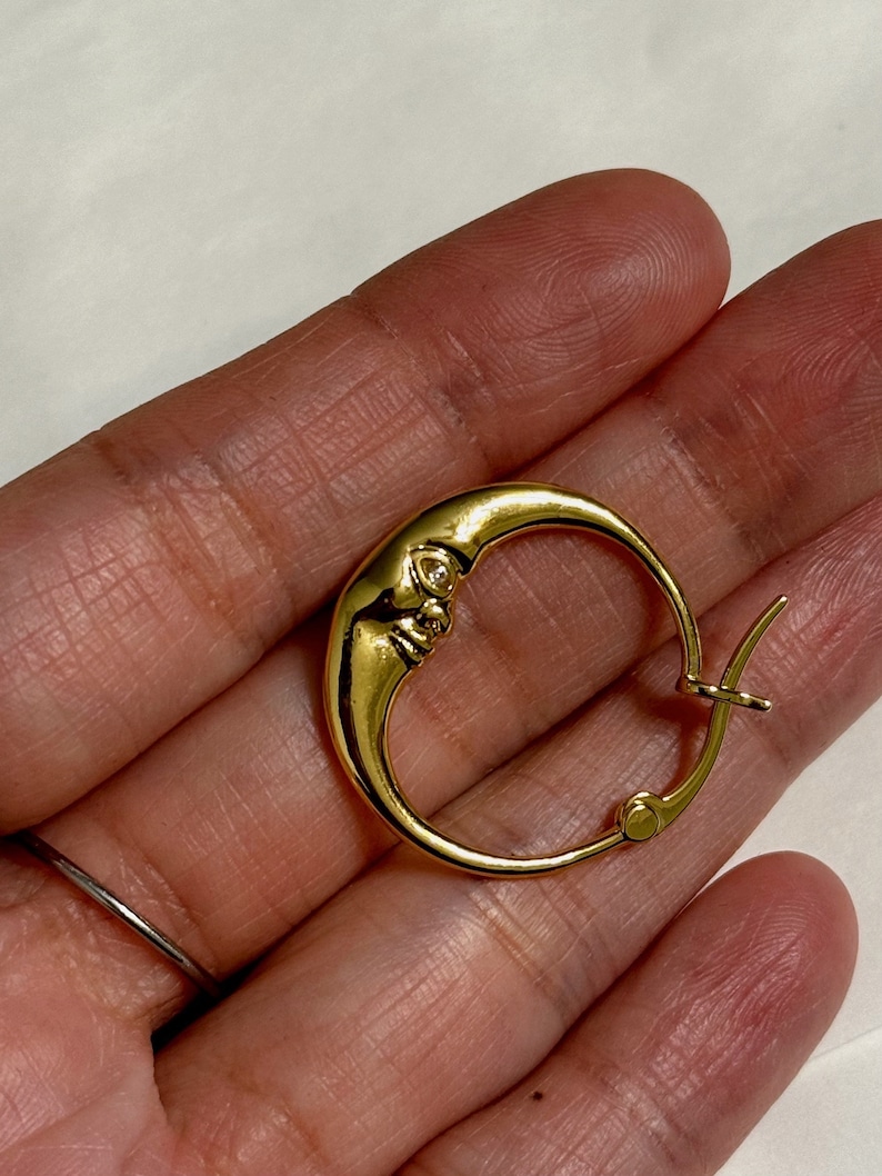 以下が含まれることがあります： 顔のデザインが施された金の三日月形のフープイヤリング。イヤリングは手で持たれています。