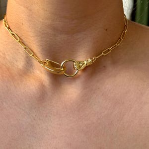 May include: Gold-toned necklace featuring a unique design. The necklace has a chain link style with a large clasp and a hand holding a ring. The necklace is a statement piece.