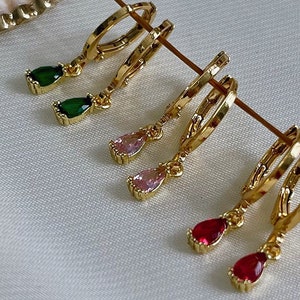 May include: Gold hoop earrings with dangling teardrop-shaped gemstones. The earrings feature green, pink, and red gemstones.
