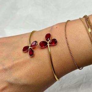 May include: Gold-toned cuff bracelet featuring two flower-shaped designs with deep red gemstones. The bracelet has a delicate, open design and is worn on a wrist, alongside other gold bracelets.