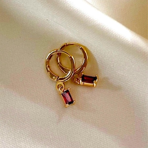 May include: A pair of gold-colored hoop earrings with rectangular, deep red gemstones dangling from the hoops. The earrings are displayed on a cream-colored, satin-like fabric.