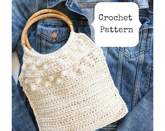 May include: A crocheted tote bag with a wooden handle. The bag is a light beige color and has a textured design. The bag is sitting on top of a blue denim jacket.