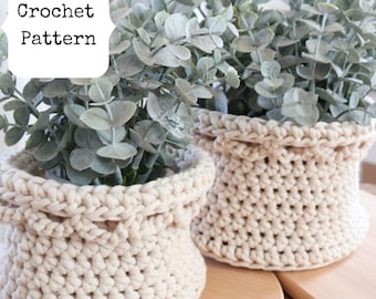 Puede incluir: Dos cestas de plantas de ganchillo de color crema con follaje verde. Las cestas son redondas con un borde superior doblado. El texto "Crochet Pattern" está en un cuadro blanco en la esquina superior izquierda. Las cestas están sobre una superficie de madera.