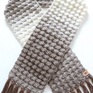 Puede incluir: Una bufanda de ganchillo con un degradado de marr&oacute;n, beige y blanco. La bufanda tiene flecos marrones en los extremos.