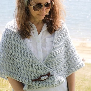 May include: A woman wearing a light gray crochet shawl with a wooden pin. The shawl is draped over her shoulders and fastened with a pin. She is also wearing a white shirt and a light gray headband.