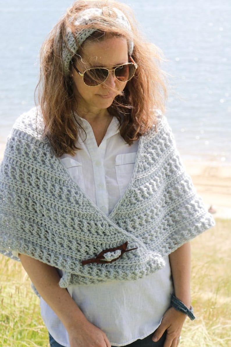 May include: A woman wearing a light gray crochet shawl with a wooden pin. The shawl is draped over her shoulders and fastened with a pin. She is also wearing a white shirt and a light gray headband.