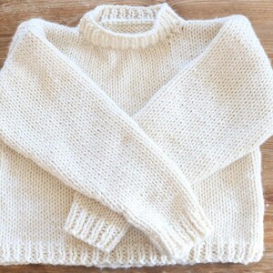 May include: Cream-colored, hand-knitted sweater with a crew neck and long sleeves. The sweater is made of a thick yarn, with ribbed cuffs and hem. The garment is displayed on a wooden surface.