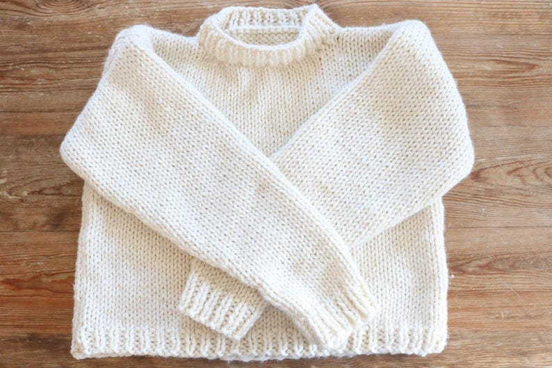 May include: Cream-colored, hand-knitted sweater with a crew neck and long sleeves. The sweater is made of a thick yarn, with ribbed cuffs and hem. The garment is displayed on a wooden surface.