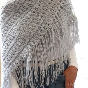 May include: A light gray crochet shawl with a fringe border. The shawl is worn by a person wearing a white shirt and blue jeans.
