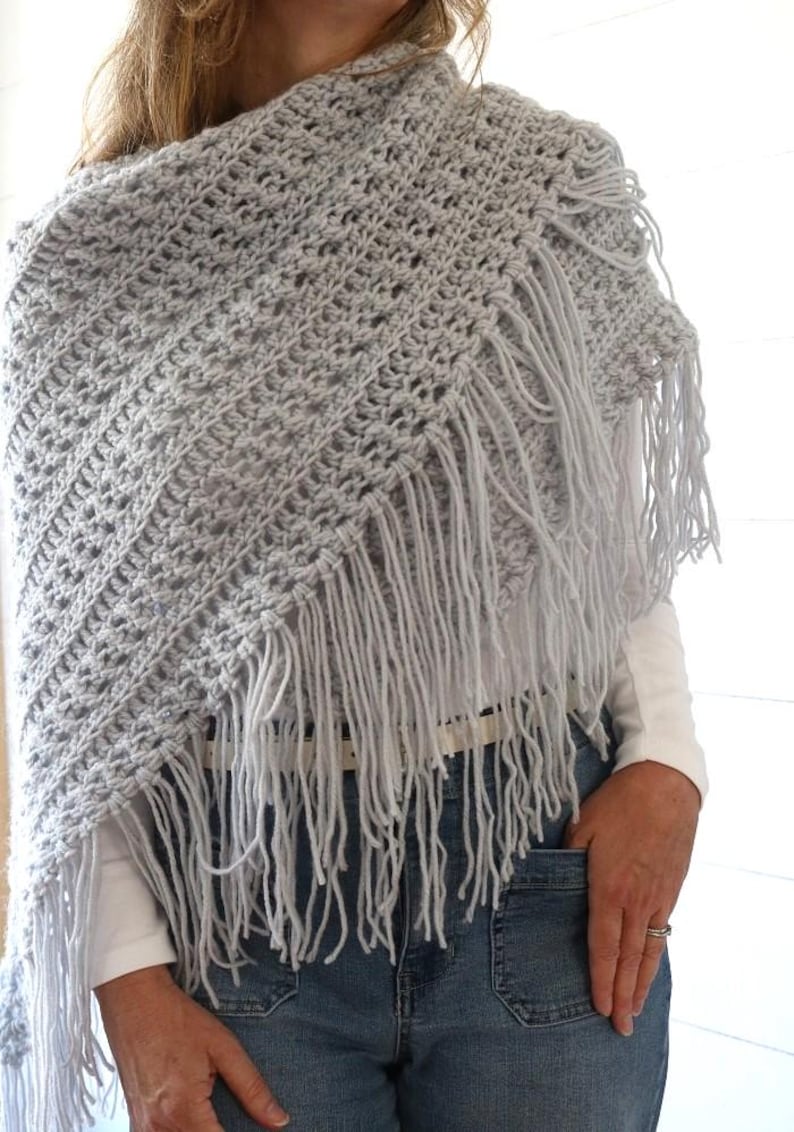 May include: A light gray crochet shawl with a fringe border. The shawl is worn by a person wearing a white shirt and blue jeans.