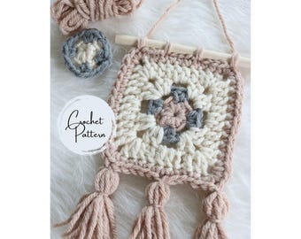 May include: A crochet wall hanging with a square design in white, pink, and gray yarn. The hanging has three tassels at the bottom and is attached to a wooden dowel.