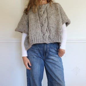 Puede incluir: Un poncho de punto gris con un dise&ntilde;o de punto trenzado en la parte delantera. El poncho se lleva sobre una camiseta blanca de manga larga y se combina con vaqueros azules. Un tapiz de macram&eacute; est&aacute; en el fondo.