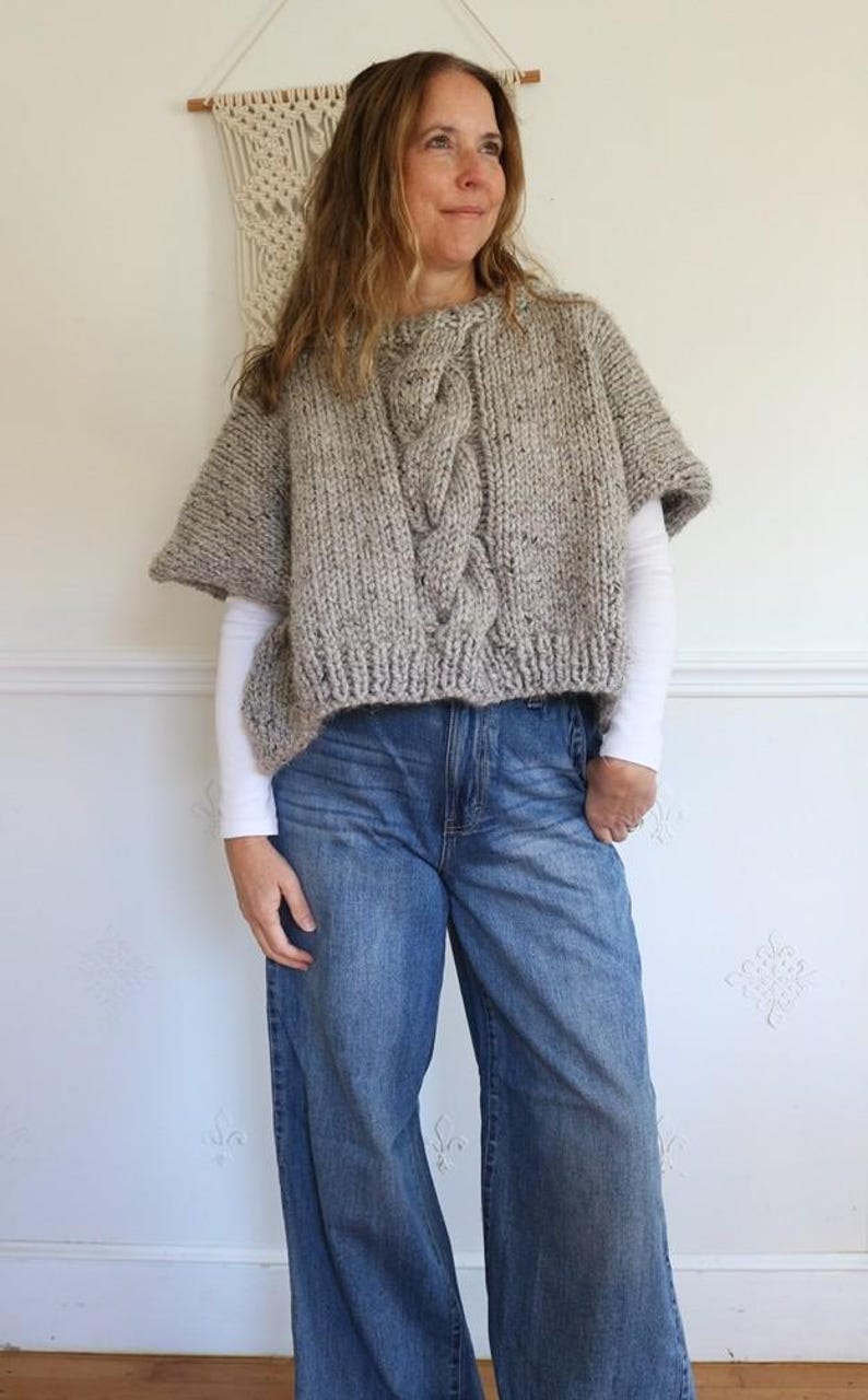 Puede incluir: Un poncho de punto gris con un dise&ntilde;o de punto trenzado en la parte delantera. El poncho se lleva sobre una camiseta blanca de manga larga y se combina con vaqueros azules. Un tapiz de macram&eacute; est&aacute; en el fondo.