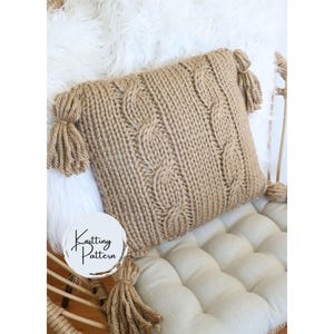 Könnte beinhalten: Ein beigefarbenes Strickkissen mit Zopfmuster und Quasten an jeder Ecke. Das quadratische Kissen scheint handgefertigt zu sein. Der Text "Knitting Pattern" befindet sich auf einem weißen Kreis in der unteren linken Ecke.