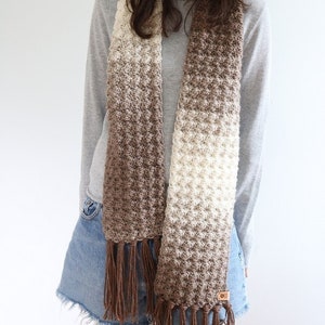 Puede incluir: Una bufanda de crochet marr&oacute;n y blanco degradado con largas borlas marrones. La bufanda la lleva puesta una persona que lleva una camisa de manga larga gris y pantalones cortos de mezclilla.