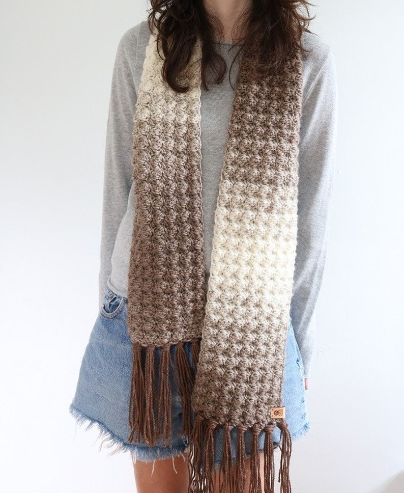 Puede incluir: Una bufanda de crochet marr&oacute;n y blanco degradado con largas borlas marrones. La bufanda la lleva puesta una persona que lleva una camisa de manga larga gris y pantalones cortos de mezclilla.