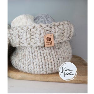 May include: A white knitted basket with a brown leather tag that says "hand made". The basket is filled with balls of yarn and is sitting on a wooden surface. Knitting Pattern.