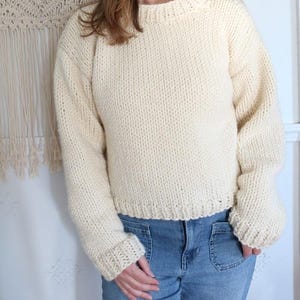 May include: Cream-colored, chunky knit sweater with a ribbed crew neck, cuffs, and hem. The long-sleeved sweater is made of a thick, textured yarn and is paired with blue jeans. The sweater has a relaxed fit.