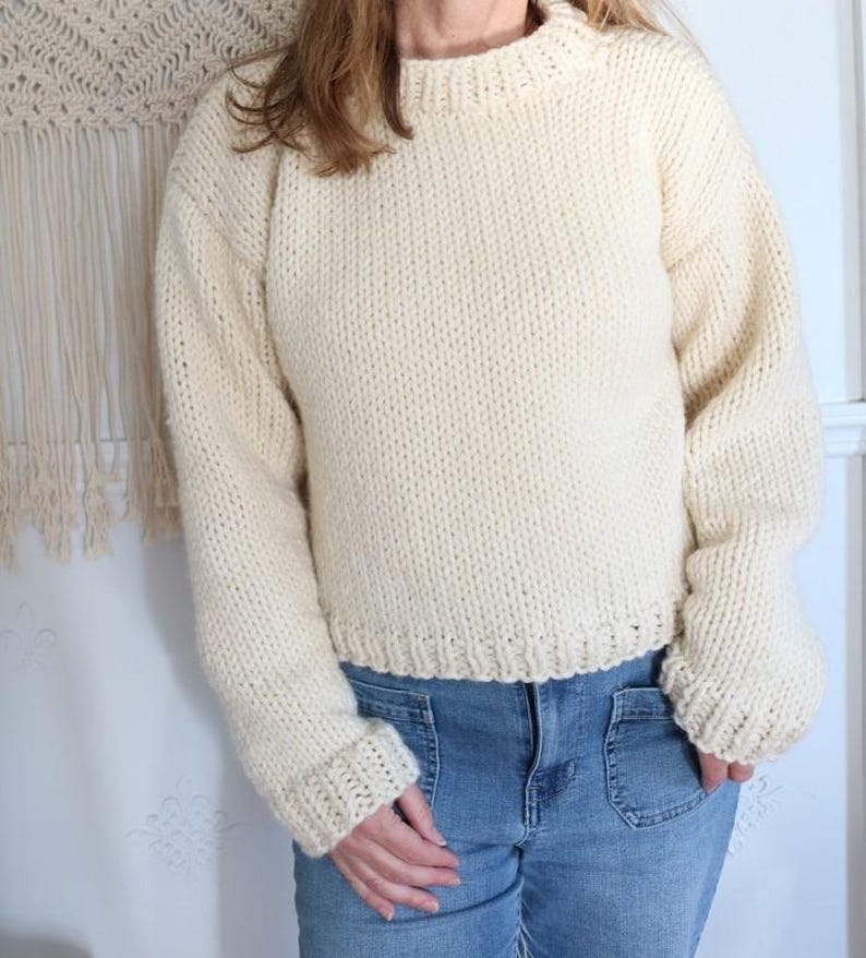 May include: Cream-colored, chunky knit sweater with a ribbed crew neck, cuffs, and hem. The long-sleeved sweater is made of a thick, textured yarn and is paired with blue jeans. The sweater has a relaxed fit.