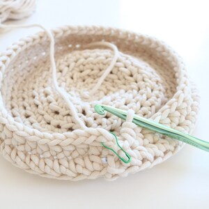 Puede incluir: Primer plano de una cesta de ganchillo de color crema en proceso. Se utiliza un ganchillo verde y un marcador de punto verde. Una madeja de hilo de color crema est&aacute; en el fondo. La cesta es redonda.