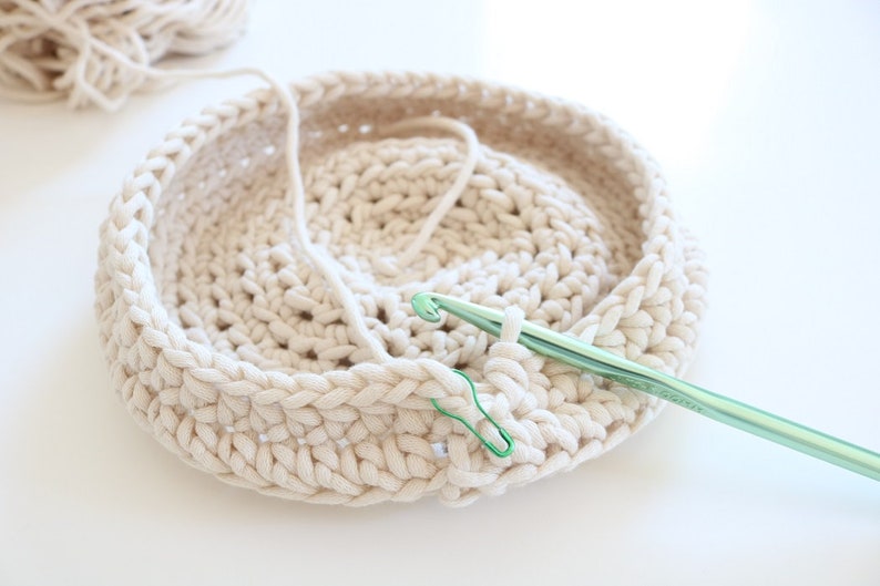 Puede incluir: Primer plano de una cesta de ganchillo de color crema en proceso. Se utiliza un ganchillo verde y un marcador de punto verde. Una madeja de hilo de color crema est&aacute; en el fondo. La cesta es redonda.