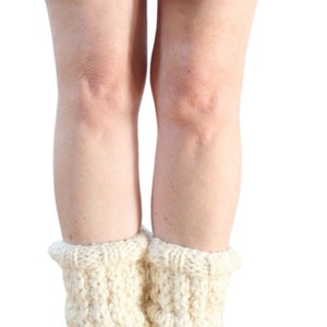 May include: A pair of white knitted leg warmers with a cable knit pattern. The leg warmers are worn over a pair of brown ankle boots.