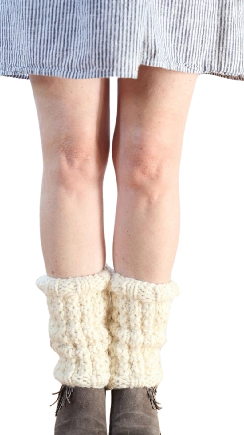 May include: A pair of white knitted leg warmers with a cable knit pattern. The leg warmers are worn over a pair of brown ankle boots.